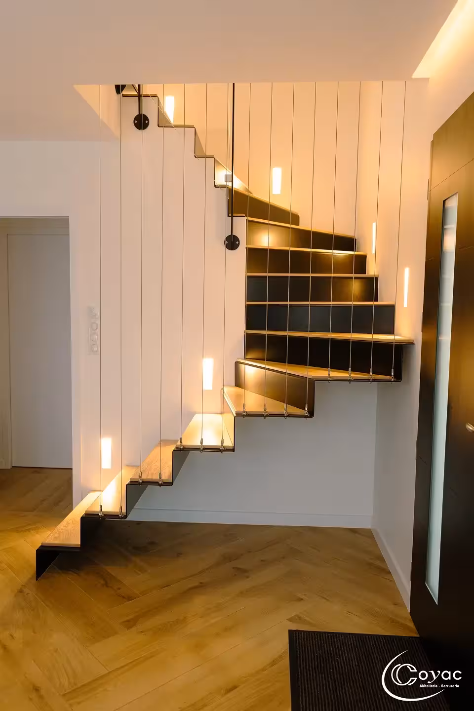 Vue d'ensemble d'un escalier sur-mesure métal et bois à Savenay au-dessus d'un parquet en chevrons, effet de suspension par câbles inox, fabriqué par Coyac